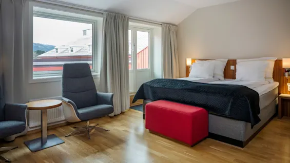 A cozy hotel room with a comfortable bed, two armchairs, and a window offering a view of buildings and mountains.