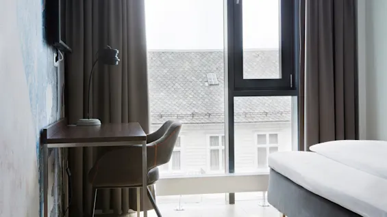 Et skrivebord med lampe og stol står ved et vindu i et minimalistisk soverom med gardiner, med utsikt over en bygning med flislagt tak.