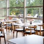 Moderni ravintola, jossa on katetut puupöydät ja tuolit suurten ikkunoiden äärellä ja näkymä vihreyteen.