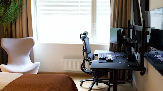 Et hotelværelse med en moderne arbejdsplads udstyret med skrivebord, ergonomisk stol og dataskærme ved vinduet