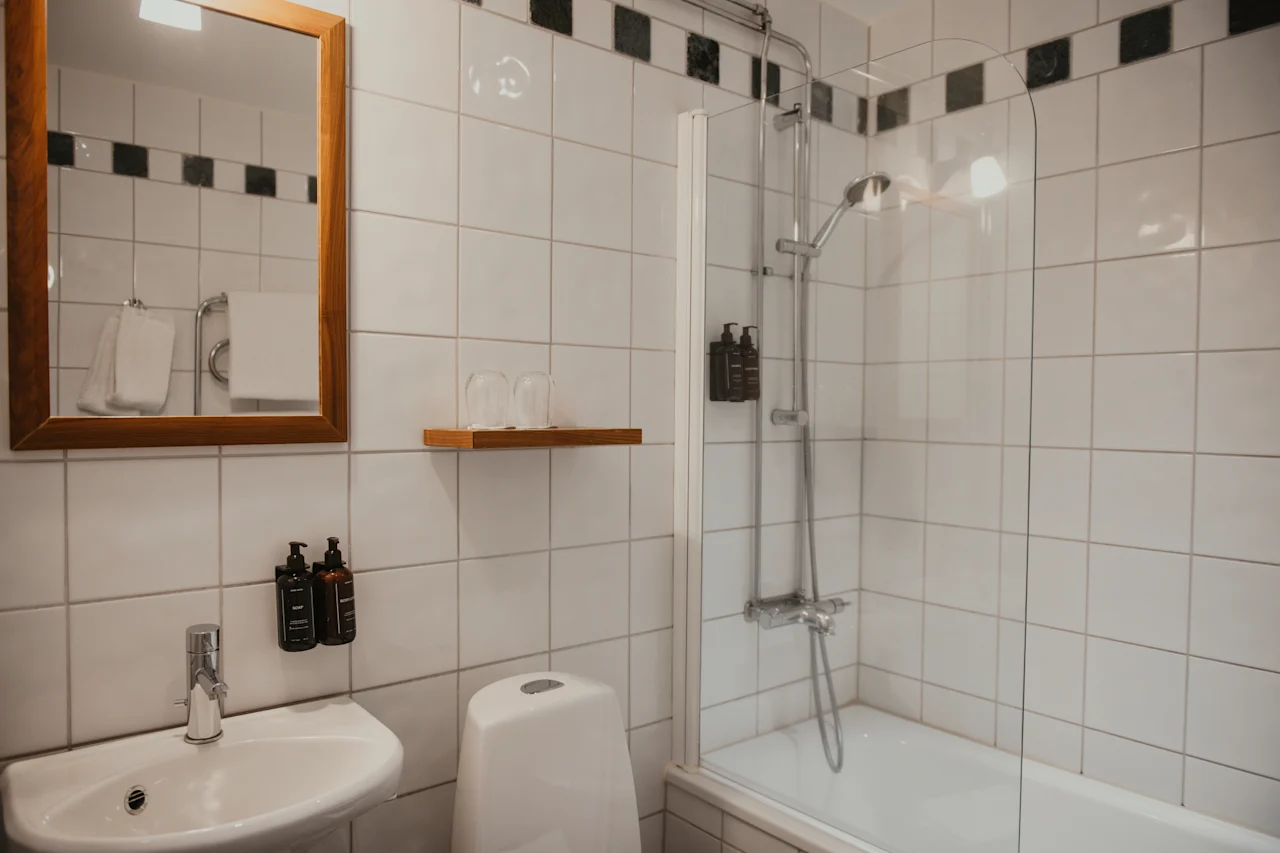 Et lyst og funksjonelt hotellbad med hvite fliser, vask, toalett og en dusj-badekar-kombinasjon.