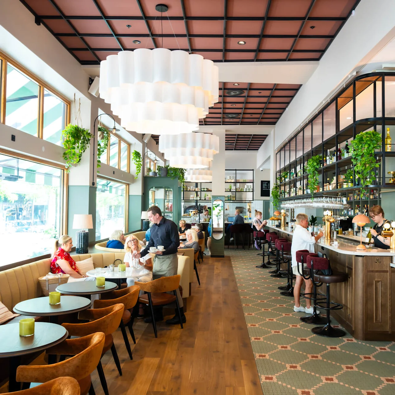 A lively cafe features people dining and conversing at tables and a bar. Large windows and hanging plants create a bright, airy atmosphere. Staff serve drinks amid stylish decor.