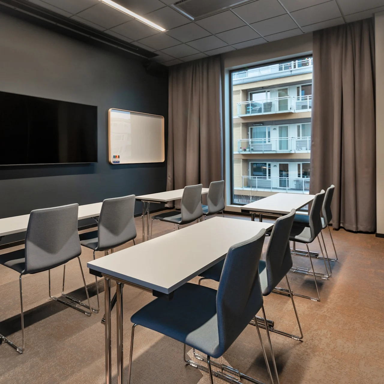Klasserom med rader av grå stoler og hvite bord vendt mot en svart tavle og TV-skjerm; utenfor viser vinduet balkonger i en nabobygning.