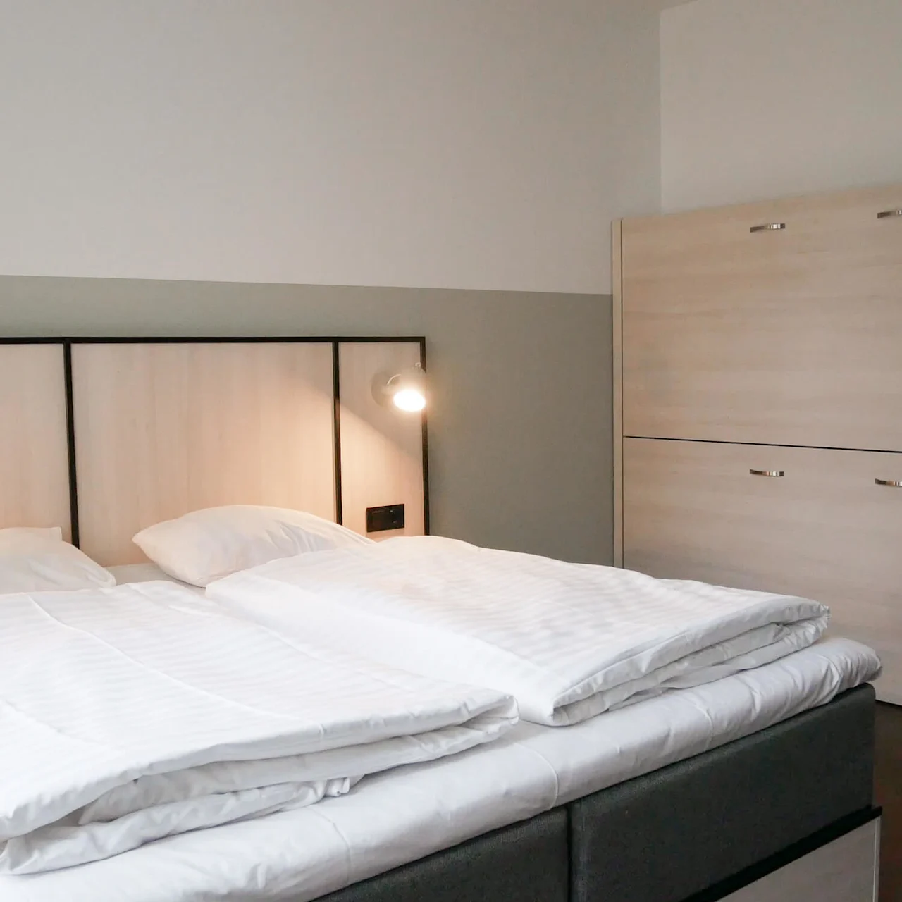 En pent oppredd seng med hvitt sengetøy står under en tent vegglampe, ved siden av et lyst treskap i et minimalistisk rom.