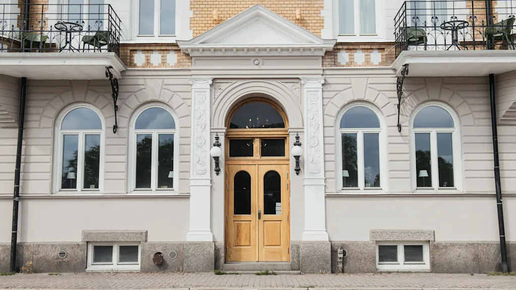 Ett par trädörrar sitter i centrum av en klassisk fasad med välvda fönster och dekorativa kolumner. Tegel- och putsbyggnaden har små balkonger och stensteg till ingången.