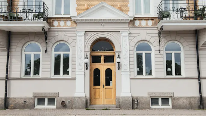 En dobbel tredør sitter i sentrum av en klassisk fasade med buede vinduer og dekorative søyler. Mur- og stukkbygningen har små balkonger og steintrinn til inngangen.