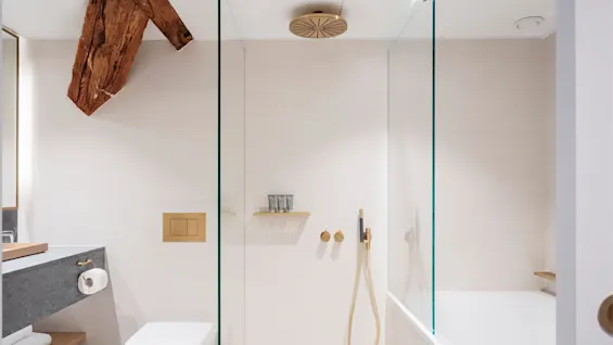 A modern bathroom features a glass-enclosed shower with gold fixtures, a white toilet, and wooden beam accents. Neutral-colored walls and a sleek vanity create a clean, minimalist environment.