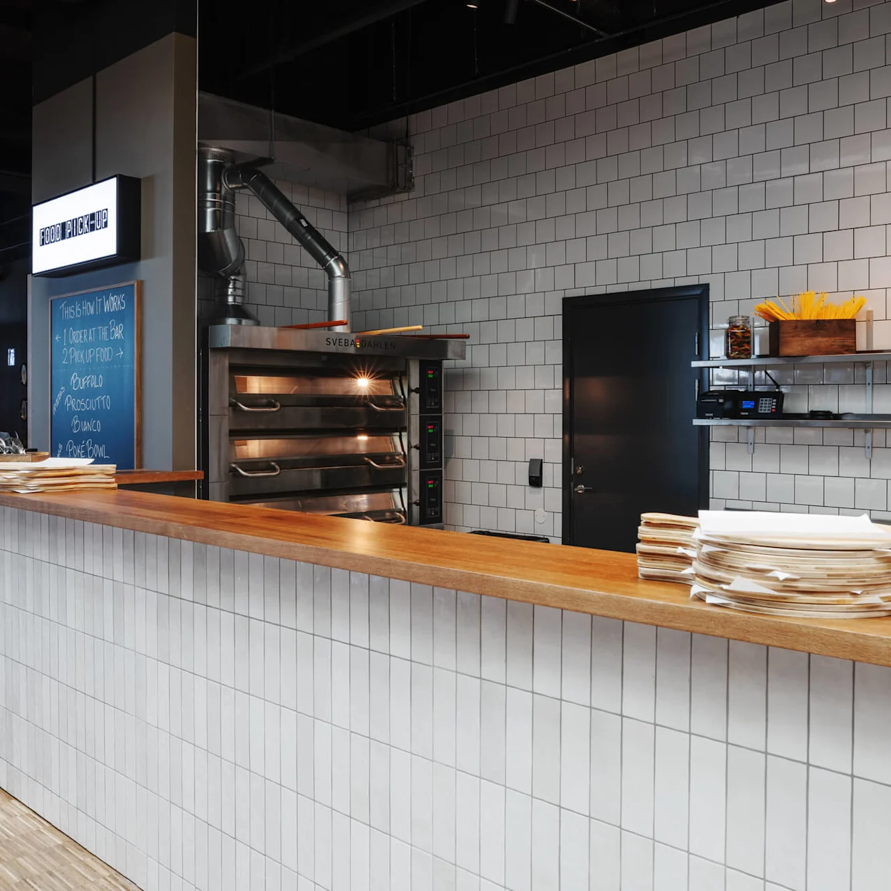 Counter with wooden top displays plates and glass jars; oven and chalkboard with instructions "1 Order at the bar, 2 Pick up food" in a modern, tiled restaurant setting.