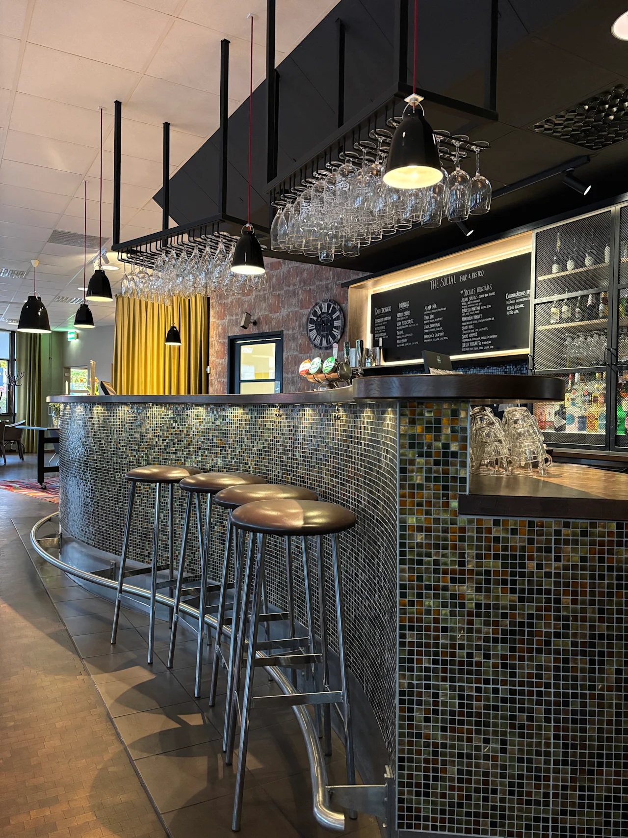 En modern bar med mosaikklädd disk, barstolar och hängande glas. En menybräda läser THE SOCIAL BAR BISTRO.
