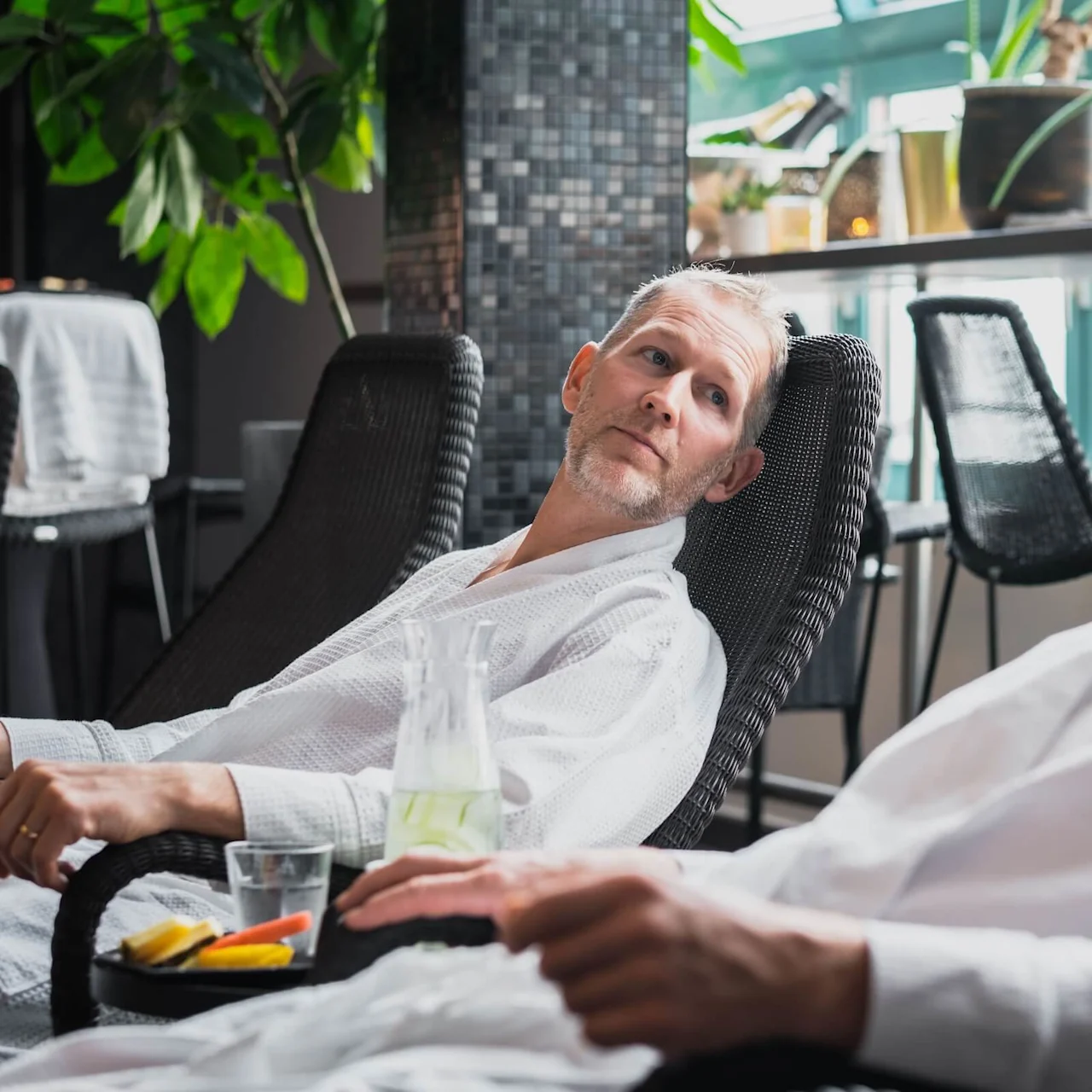 Two men, wearing white robes, lounge on wicker chairs in a bright room with large windows. A table nearby holds snacks, a glass, and a jug of water with lemon.