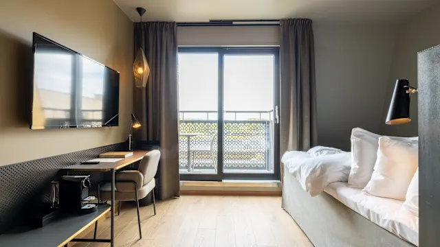 A modern hotel room with a desk, chair, bed, and balcony door, bathed in natural light.