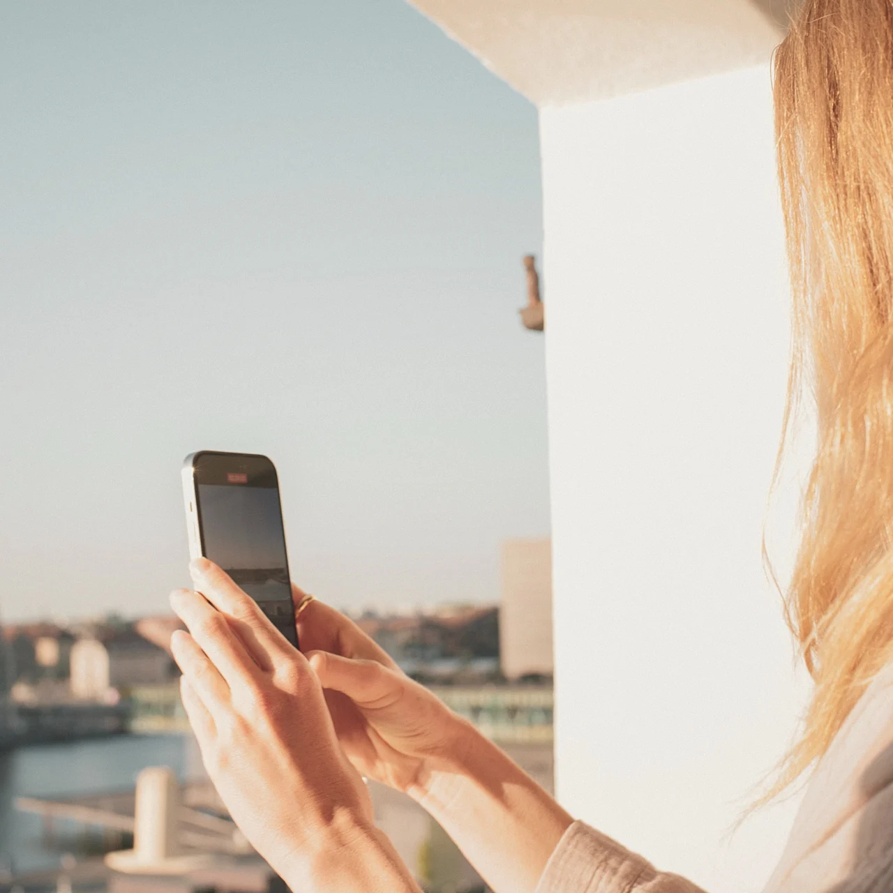 A person with long hair holds a smartphone facing a sunny cityscape from a balcony. Modern buildings and water are visible in the background.