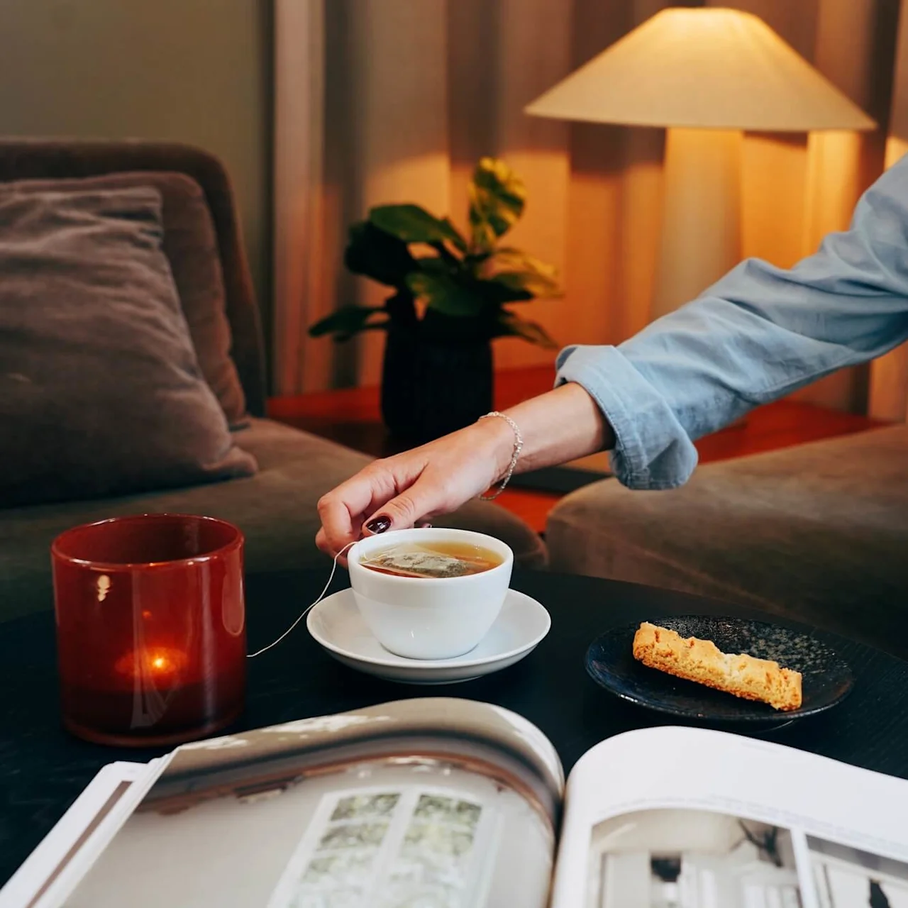 En person rör om i en kopp te på ett svart bord, med ett ljus, biscotti och en tidning, i en mysig inomhusmiljö.