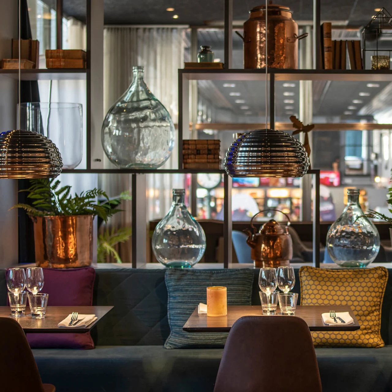 Tre bord med glas och servetter i en mysig restaurang, omgivna av dekorativa glasflaskor, kopparkannor och växter, under metallhänglampor.