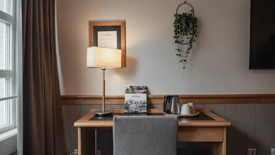 A cozy desk area in a room with a lamp, chair, and coffee/tea setup, next to a window with curtains.