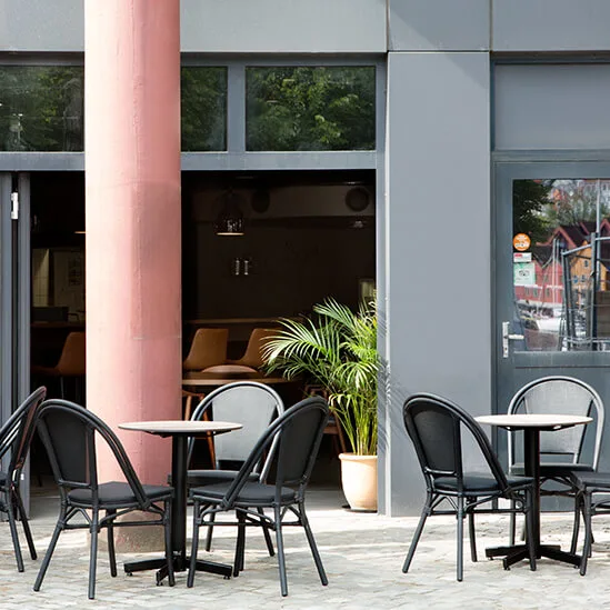 Udendørs caféborde med sorte stole er arrangeret på en brostensbelægning. En rød murstensvæg og en stor søjle danner baggrunden med en døråbning indad.