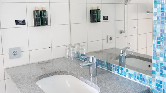 Ett handfat med en silverkran är placerat på en grå bänkskiva, omgiven av vita kaklade väggar och blå mosaikdetaljer. Monterade dispensrar innehåller toalettartiklar. Text: "WANT TO SHARE; Clarity saves the future."