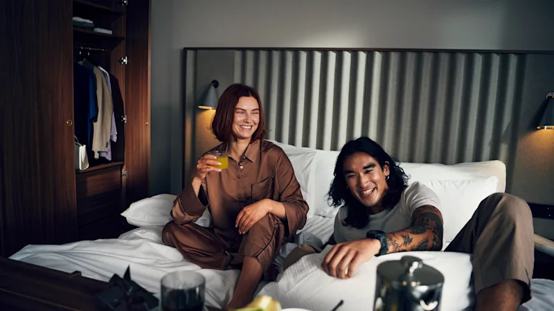 To personer nyder morgenmad på sengen, griner og deler et hyggeligt øjeblik på et hotelværelse.