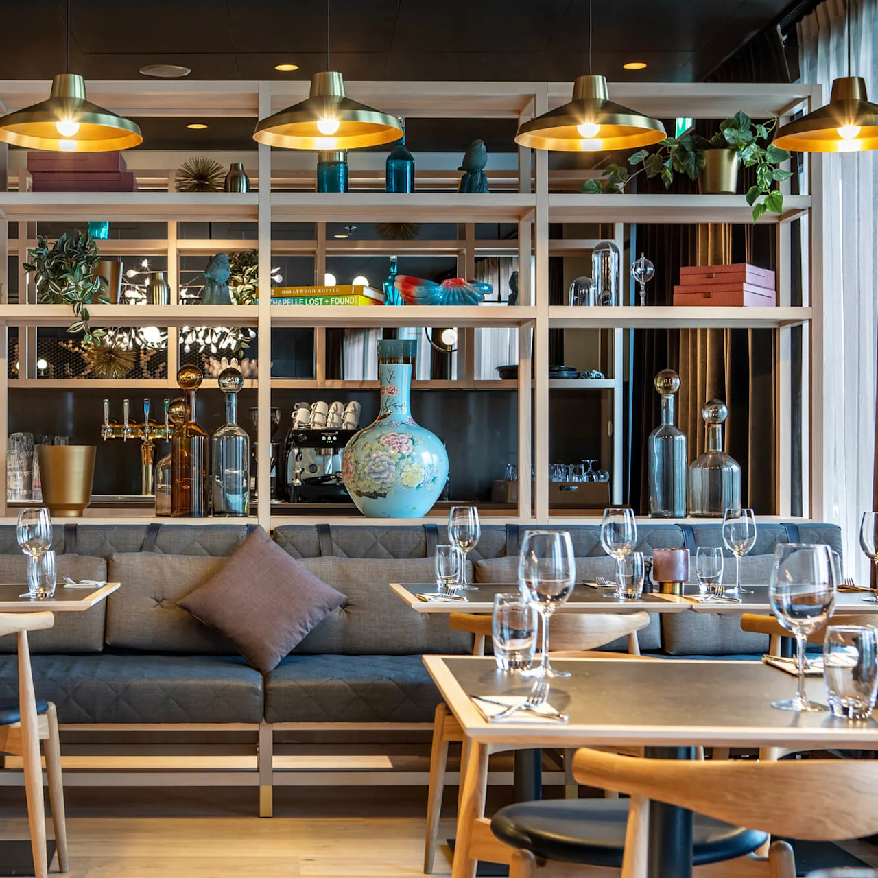 Tables set with glasses and napkins in a cozy restaurant, framed by wooden shelves holding decorative blue vases and books. Soft lighting from hanging lamps creates a warm atmosphere.