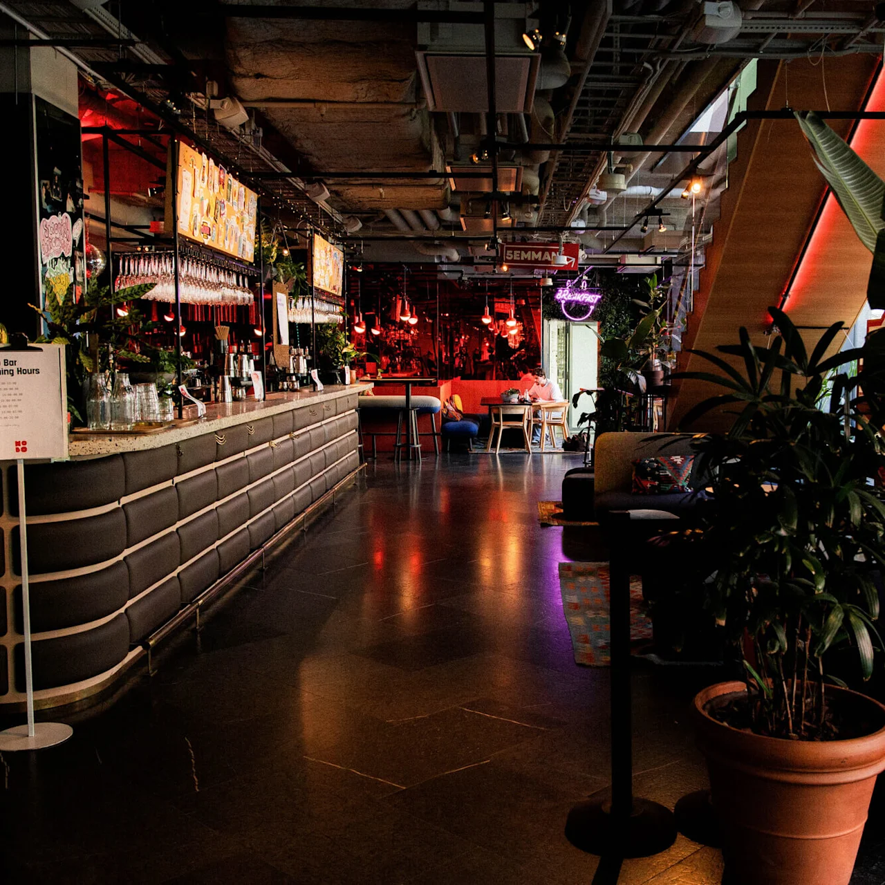 Bar with stools and plants under a ceiling. People sit in a relaxed atmosphere with ambient lighting. Sign reads "Hobo Bar Opening Hours" with timings.