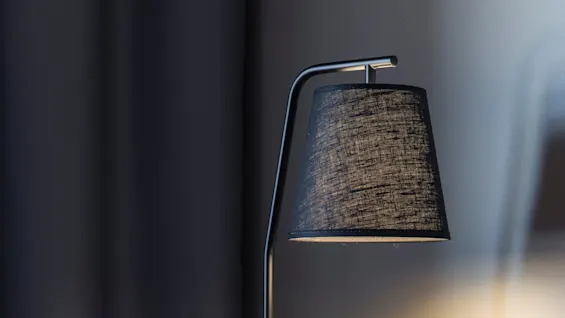En svart skrivbordslampa kastar ljus på ett träbord bredvid en röd stoppad stol, mot en bakgrund av mörka gardiner och en mjukt belyst vägg.