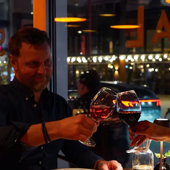 En leende man skålar med rödvinsglas med sitt sällskap vid ett mysigt restaurangbord.