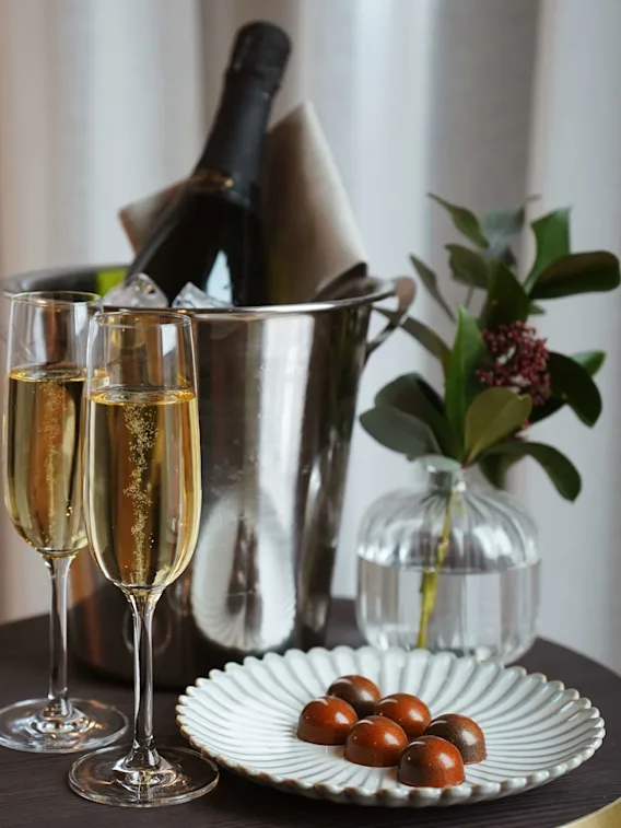 Två champagneglas med bubblor bredvid en ishink med en flaska, på ett bord med choklad, intill gardiner.