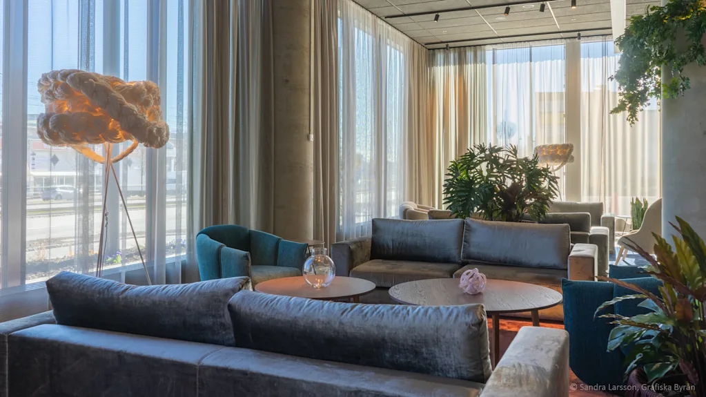 En lounge med grå soffor och runda bord står vid stora fönster med genomskinliga gardiner, belyst av naturligt ljus. En unik, vävd golvlampa står i bakgrunden.