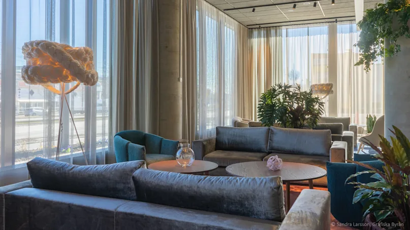 A lounge area with gray sofas and round tables sits under large windows with sheer curtains, illuminated by natural light. A unique, woven floor lamp stands in the background.
