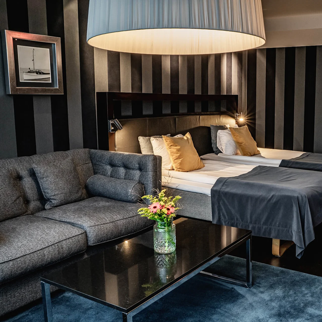 A modern hotel room contains two beds with dark linens, a gray sofa, and a black coffee table with flowers. A large lamp hangs above; striped wallpaper and a closed door are visible.
