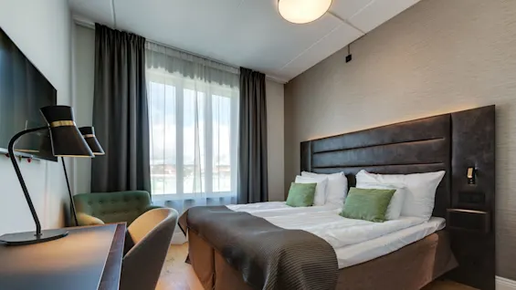Et godt opplyst hotellrom med en stor seng, stoler, et skrivebord med en lampe og gardiner som reflekterer et moderne, komfortabelt interiør.