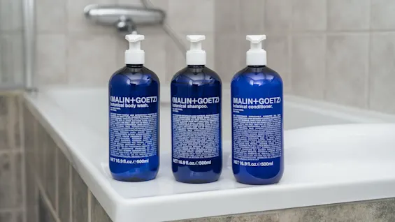 Blue bottles labeled “(MALIN+GOETZ) botanical body wash,” “botanical shampoo,” and “botanical conditioner” sit on a bathtub edge. Each bottle contains 16.9 fl. oz./500 ml. The background is a tiled bathroom wall.