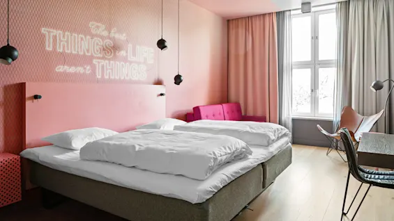 Et koselig soverom med en dobbeltseng, rosa vegger og en rosa sofa under et stort vindu. Et sitat på veggen lyder, "The best THINGS in LIFE aren’t THINGS."
