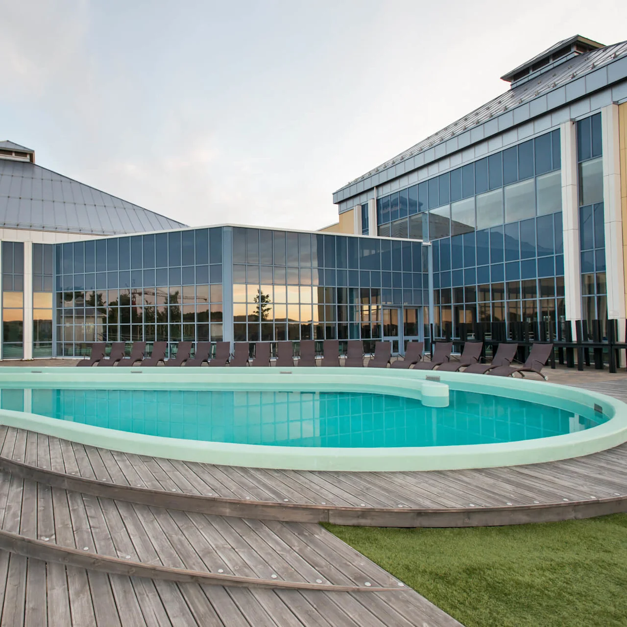 A circular outdoor swimming pool is surrounded by a wooden deck, with loungers positioned around it. The pool is adjacent to a large modern building with glass panels.