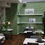 Bord och stolar är arrangerade i ett modernt, växtfyllt café med gröna väggar och inramade bilder. Miljön har trägolv, vilket ger en mysig atmosfär.