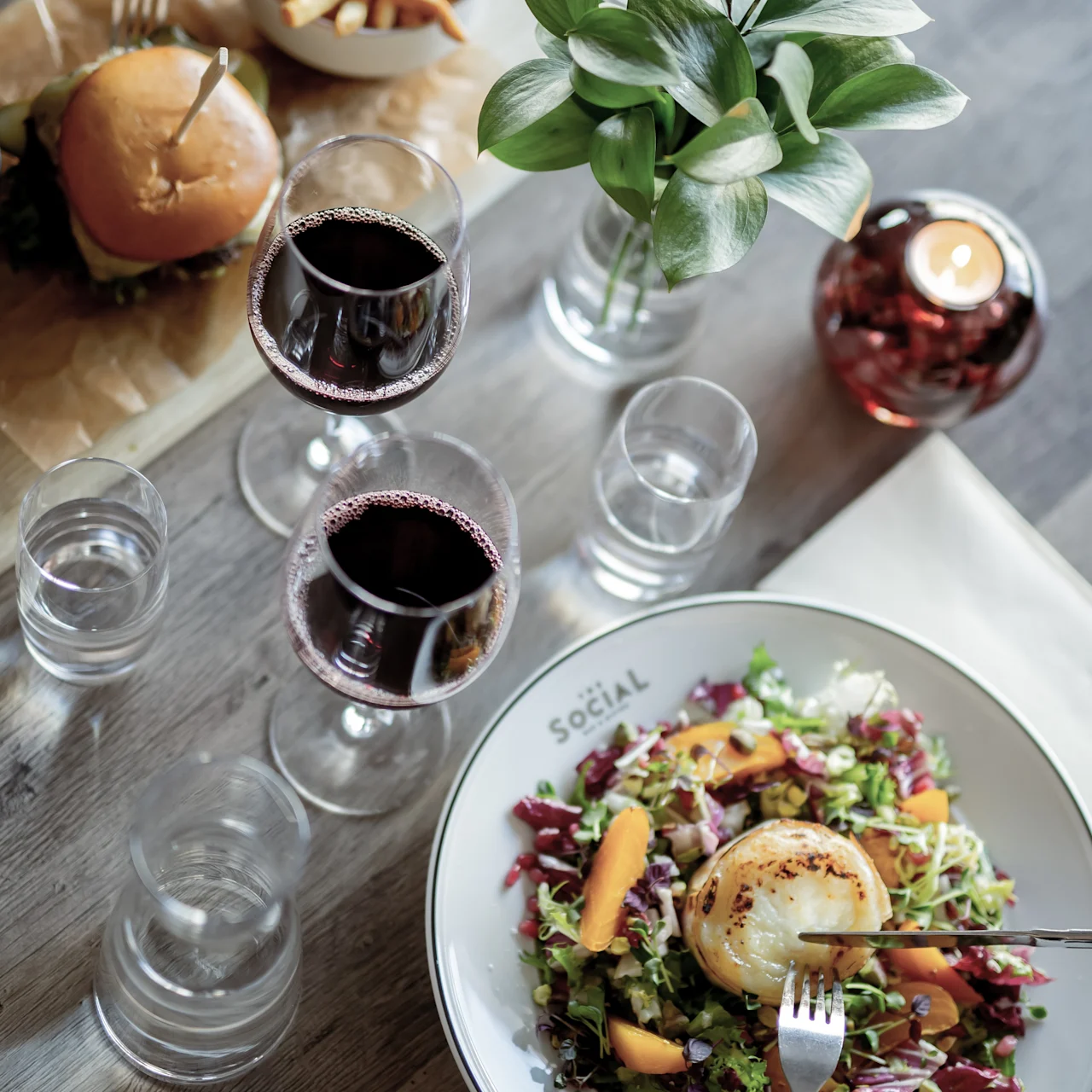 A salad with grilled cheese is being eaten on a table alongside a burger, fries, wine glasses, and a small plant. The plate reads "SOCIAL."