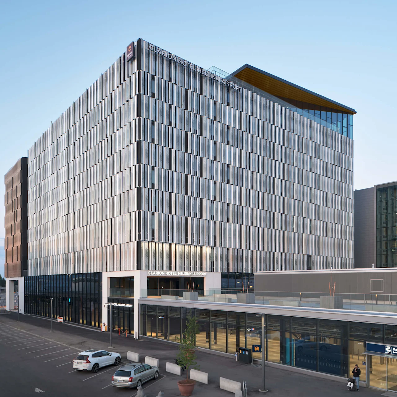 A modern, glass-clad hotel building with "Clarion Hotel Helsinki Airport" signage; situated next to a railway station. Two parked cars and a clear blue sky create a calm urban scene.
