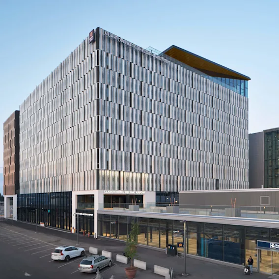 Nykyaikainen, lasiseinäinen hotellirakennus kyltillä "Clarion Hotel Helsinki Airport"; vieressä rautatieasema. Kaksi pysäköityä autoa ja kirkas sininen taivas luovat rauhallisen kaupunkinäkymän.