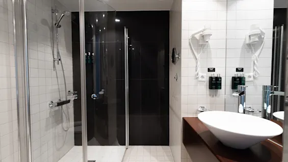 A modern bathroom features a glass-enclosed shower, wall-mounted showerhead, sink on a wooden countertop, white tiled walls, and a hairdryer mounted on the wall.