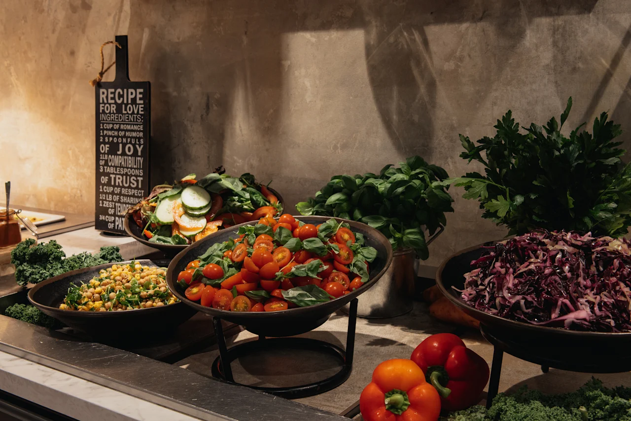 En farverig buffet med friske salater inklusive tomater, kikærter og kål, med et skilt der læser RECIPE FOR LOVE INGREDIENTS.