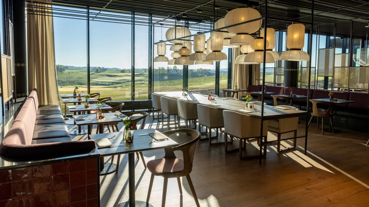 Modern restaurant interior with elegant tables and chairs; large windows provide a view of a golf course in bright daylight.