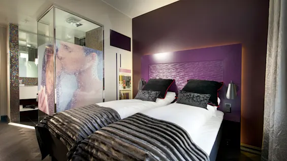 A modern hotel room features two neatly made beds with black cushions and metallic bedside lamps. A glass shower with a frosted image of a couple kissing is adjacent.
