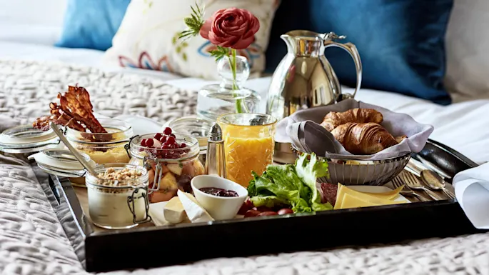 Et brett med croissanter, frukt, bacon, ost, en blomsterkrukke og et glass appelsinjuice på en seng. Dekorative puter og et teppe gir en koselig bakgrunn.