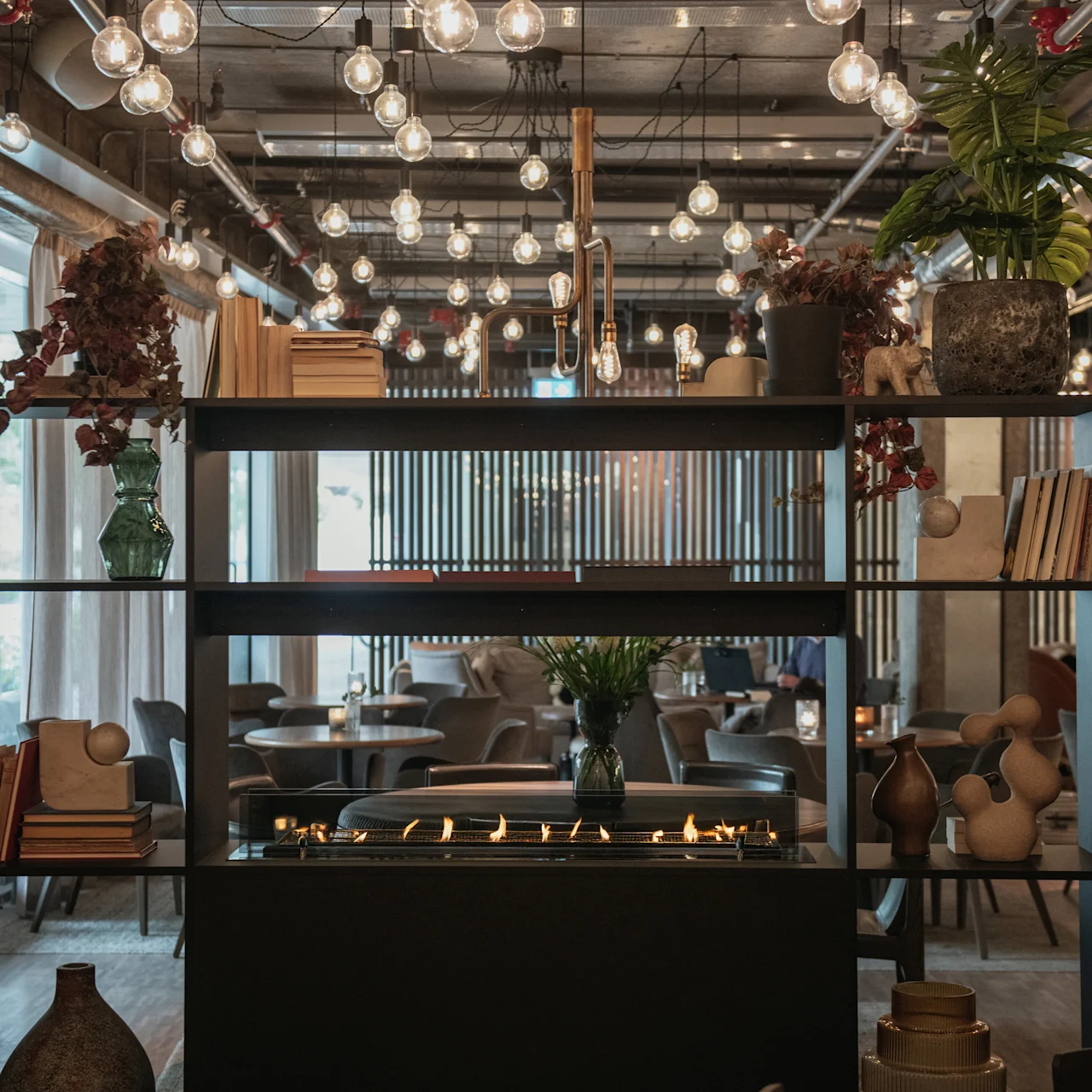 A shelf with books and plants stands in a cozy restaurant, featuring a central fireplace below. Soft lighting with numerous hanging bulbs creates an inviting ambiance, surrounded by tables and chairs.