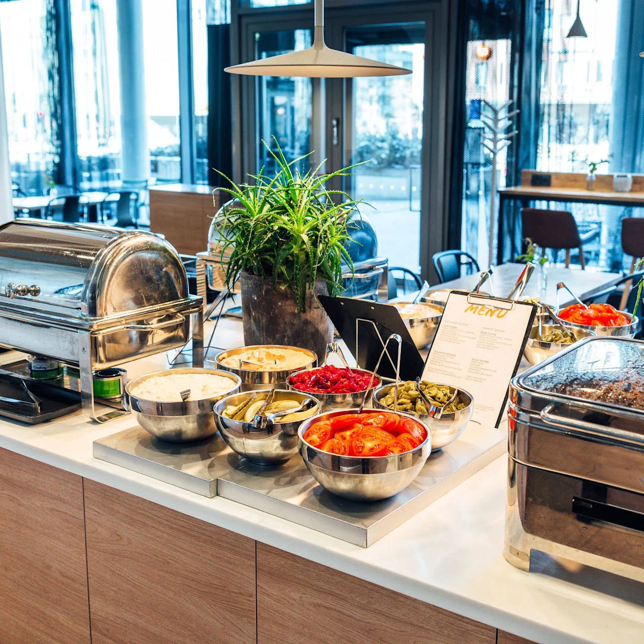 Buffet setup with various food bowls containing pickles, tomatoes, and other items on a counter. Large chafing dishes are present. Indoor dining space visible with tables and chairs. Menu sign visible.