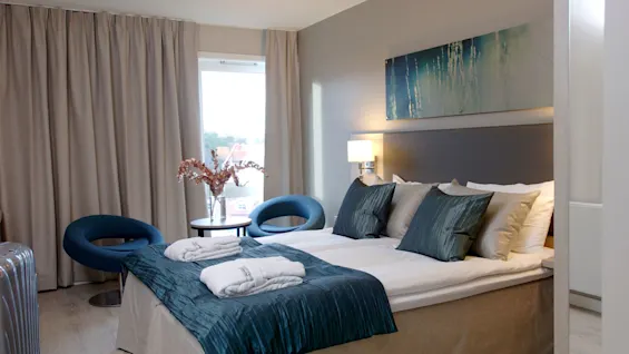 En seng med teal-fargede puter omgitt av moderne møbler og kunst. Beige gardiner rammer inn et vindu, og morgenkåper ligger brettet på sengen.