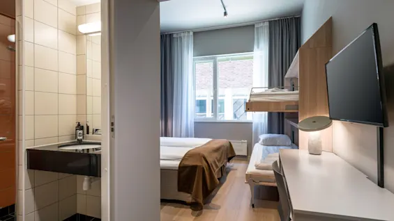 A hotel room with a double bed and a bunk bed. A desk holds a lamp and TV, while a bathroom sink is visible in an adjacent tiled room.