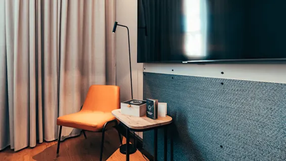 En oransje stol står i et lyst rom ved siden av et lite bord med bøker og en lampe, foran en stor mørk TV-skjerm.