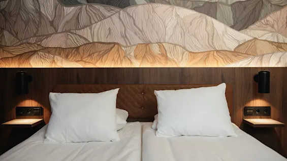 Et koselig hotellrom med en pent oppredd seng, hvite puter og en dekorativ sengegavl med abstrakt fjellignende kunst.