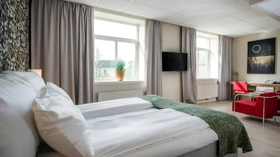 En pent oppredd seng vender mot store vinduer med grå gardiner, ved siden av en veggmontert TV. Røde stoler og et skrivebord står i hjørnet under et innrammet abstrakt kunstverk.